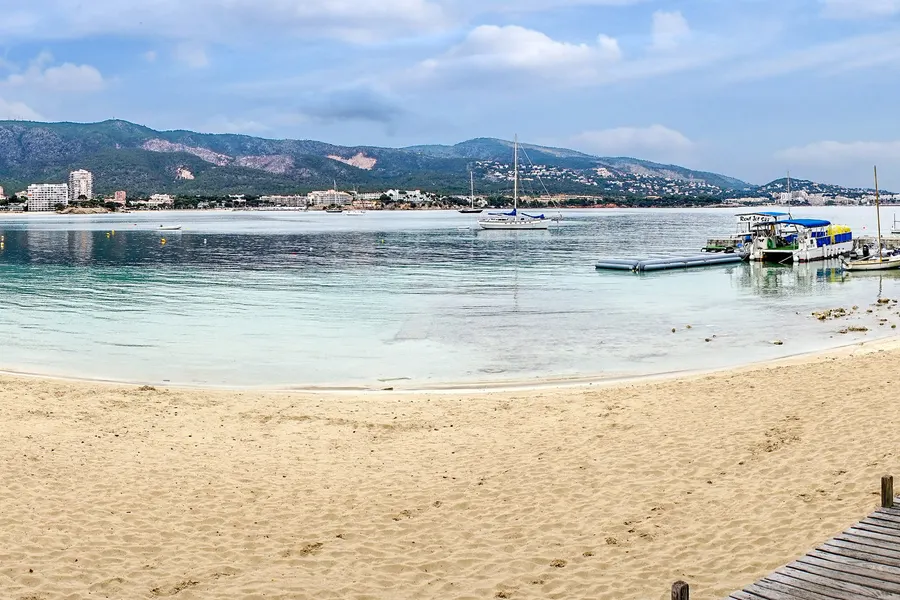 Die Bucht von Palmanova mit hellem Sand, ruhigem Wasser, einem Steg mit Booten und den bewaldeten Bergen im Hintergrund.
