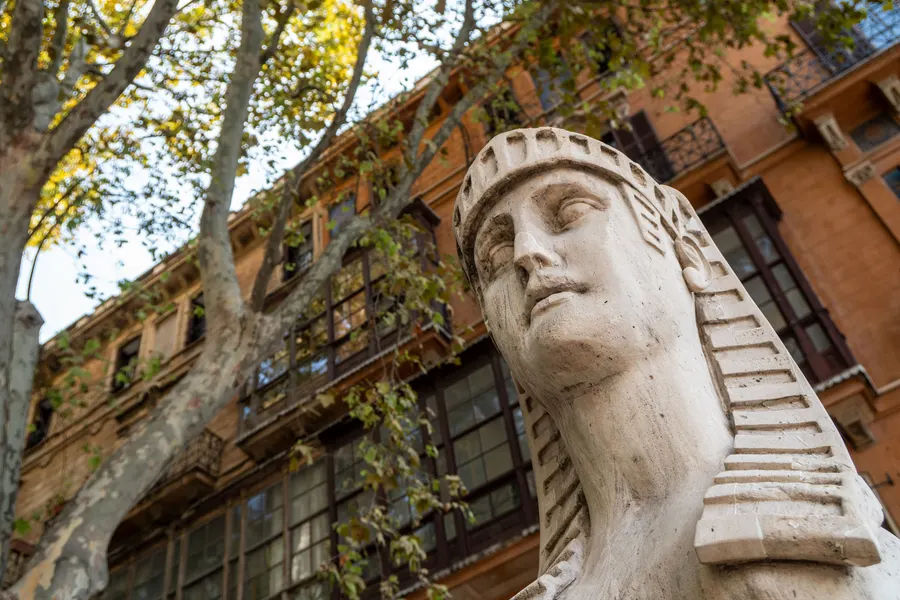 Nahaufnahme einer steinernen Sphinx am Passeig del Born in Palma, dahinter eine Fassade mit Balkonen und Platanenäste.
