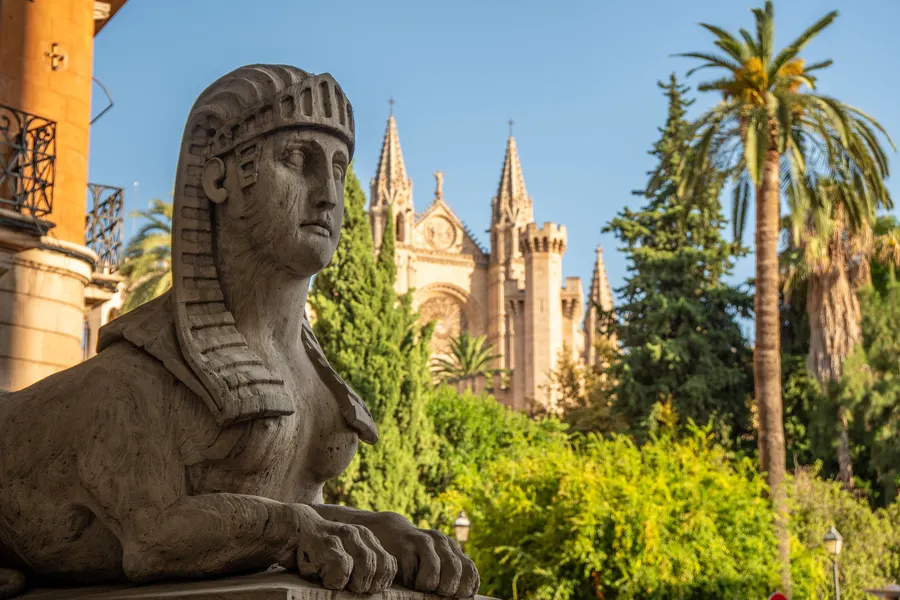 Steinerne Sphinx im Profil vor Palmen, im Hintergrund die Türme der Kathedrale von Palma.
