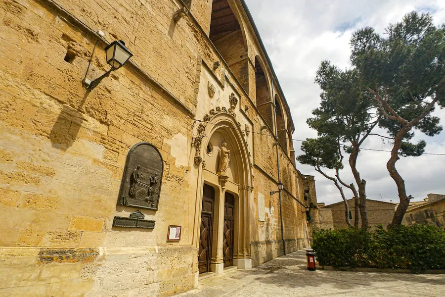 Seitenfassade der Klosterkirche Sant Bernardí in Petra mit Rundbogenportal, Reliefnische und Gedenktafel, daneben Kiefern.