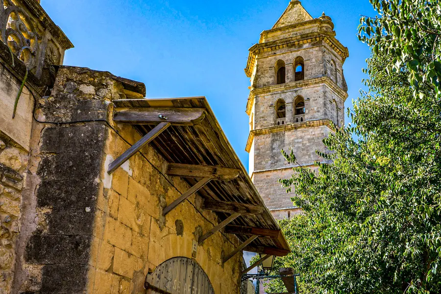 Der Glockenturm der Pfarrkirche von Petra über den Dächern, davor ein altes Wirtschaftsgebäude mit hölzernem Vordach und ein Feigenbaum.