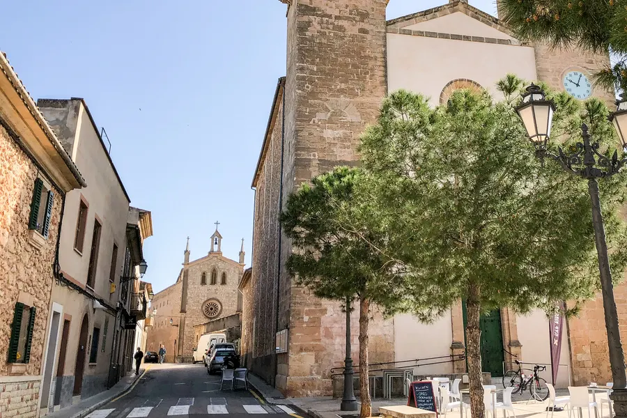 Straße in Pina mit Bäumen und niedrigen Häusern, am Ende der Blick auf die Kirche unter blauem Himmel.
