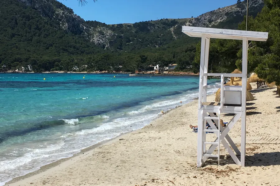 Weißer hölzerner Rettungsschwimmerturm am Strand von Formentor, davor auslaufende Wellen und einzelne Badegäste, im Hintergrund bewaldete Berge.