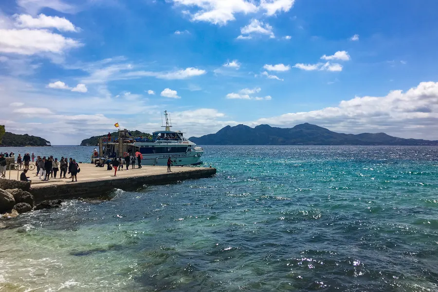Steinerner Anleger an der Platja de Formentor mit einem blau-weißen Ausflugsboot, wartenden Fahrgästen und dem offenen Wasser der Bucht dahinter.