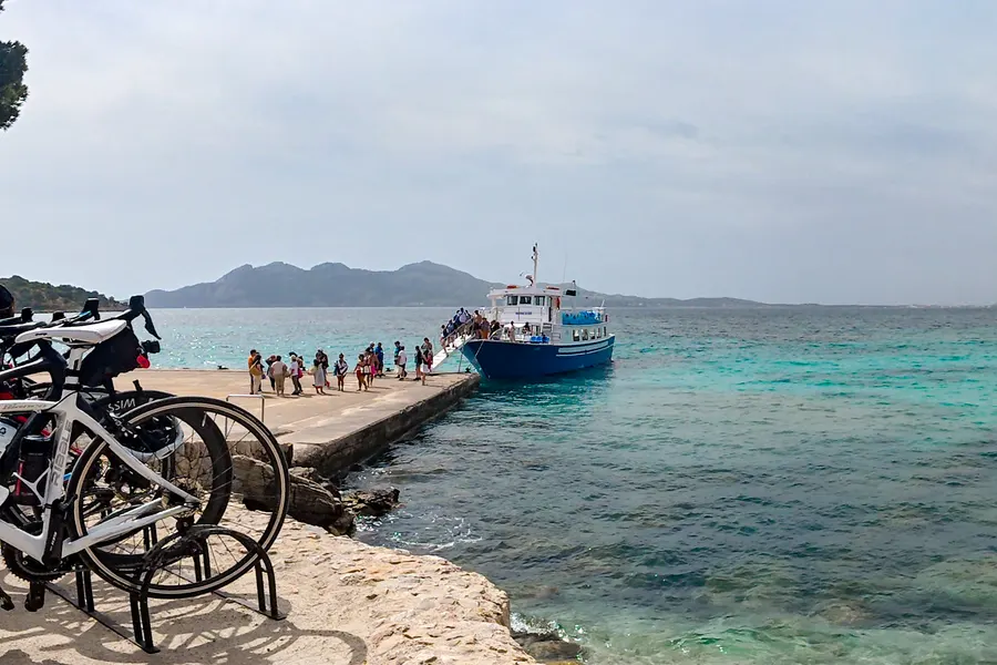 Menschenschlange auf dem Anleger vor einem anlegenden blauen Ausflugsboot, im Vordergrund abgestellte Rennräder, dahinter die Bucht.

