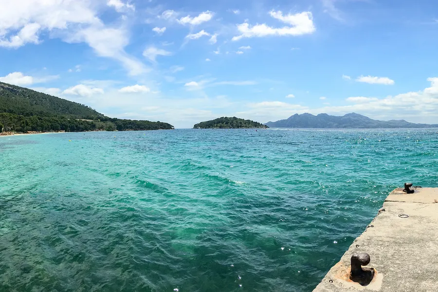 Blick vom Anleger über das türkis- und grünblau schimmernde Wasser der Bucht von Formentor auf die vorgelagerte Halbinsel und die Bergkette am Horizont.
