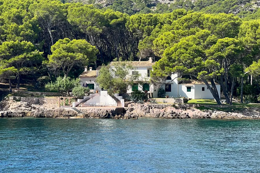 Weiße Gebäude zwischen alten Kiefern direkt am felsigen Ufer der Bucht von Formentor, davor ruhiges dunkelblaues Wasser.
