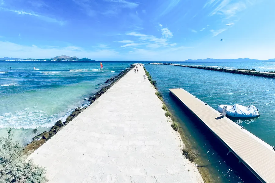 Breite gepflasterte Mole zieht sich geradeaus ins Meer und trennt links das offene, windbewegte Wasser mit Surfern vom ruhigen Kanal rechts mit Steg und abgedecktem Boot; am Horizont Berge unter blauem Himmel.