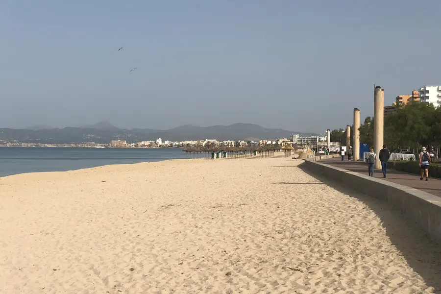Der breite helle Sandstrand der Platja de Palma mit der Promenade rechts, dahinter die Bucht von Palma und die Berge der Tramuntana.