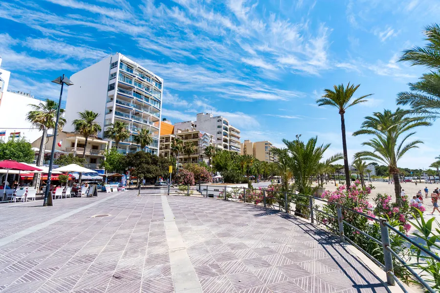 Eine steinerne Buhne, die vom Strand der Platja de Palma ins flache Wasser führt, dahinter die Promenade und die Bucht.
