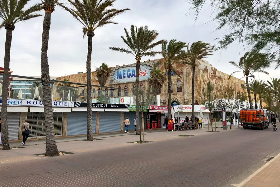 Weißes Gebäude mit Rundbögen und Turm an der Promenade der Platja de Palma, davor Bäume mit Früchten.
