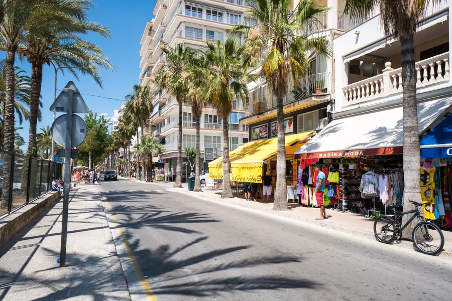 Die Strandpromenade von S'Arenal mit Geschäften, Palmen und Fahrradfahrern, mehrsprachige Beschilderung an den Fassaden.