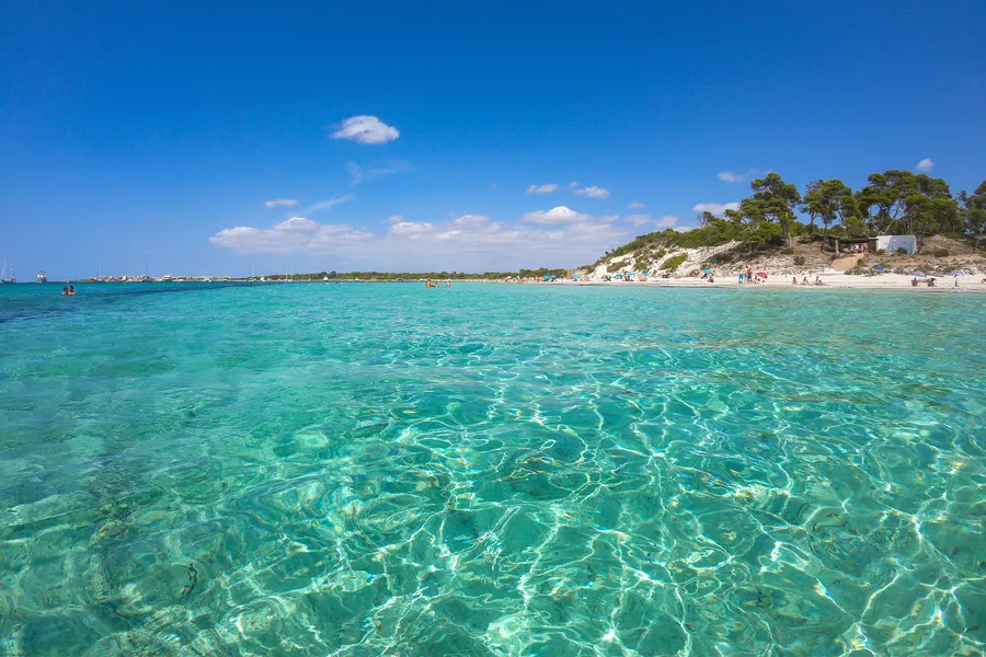 Breiter weißer Sandstrand der Platja des Carbó mit einzelnen Badegästen, im Wasser ankernde Boote und ein Rettungsring.