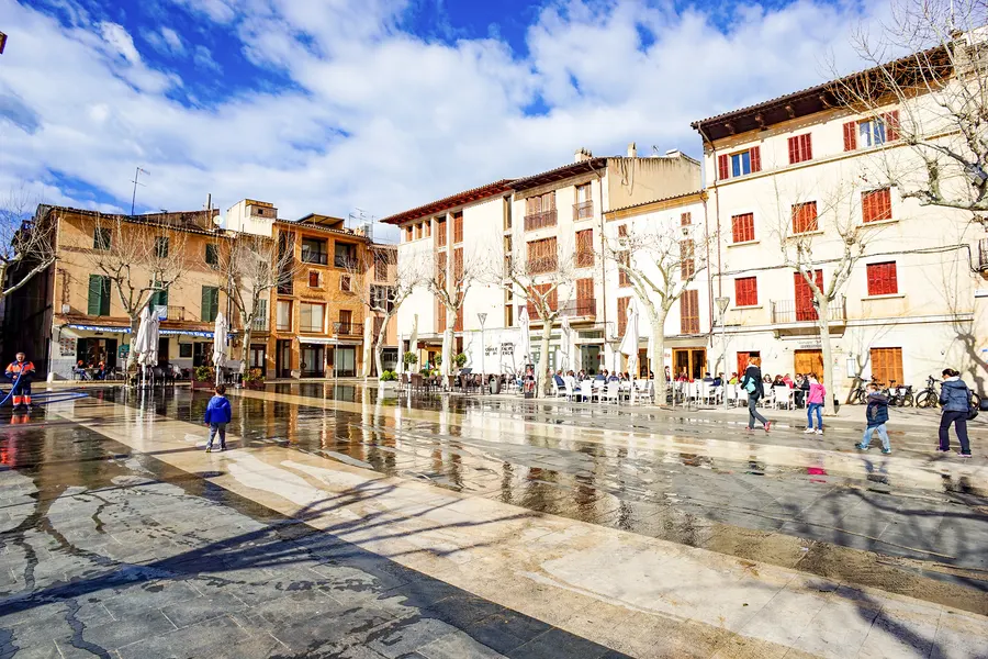 Dieselbe Plaça Major im Winter: nasses, spiegelndes Pflaster nach der Reinigung, kahle Platanen, wenige Menschen in Winterjacken, ein Kind läuft über den Platz; die Cafés unter geschlossenen Schirmen sind besetzt.