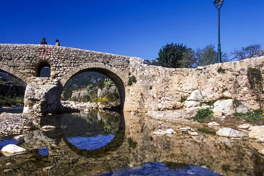 Zweibogige Bruchsteinbrücke über einen fast stillstehenden Bach, in dem sich die Bögen spiegeln; auf der Brücke zwei Personen, im Hintergrund bewaldete Berghänge unter tiefblauem Himmel.
