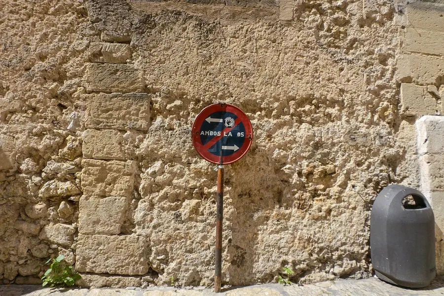 Verrostetes, ausgebleichtes Halteverbotsschild mit kaum noch lesbarer Aufschrift vor einer groben Bruchsteinmauer, rechts ein grauer Abfallbehälter auf dem Bürgersteig.
