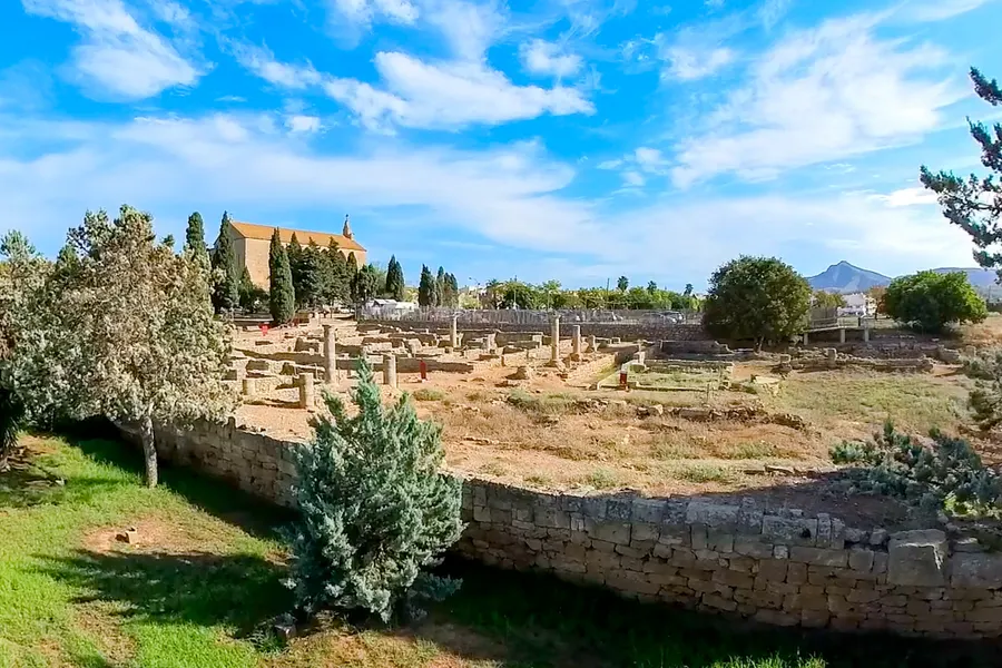 Weiter Blick über das Ausgrabungsfeld von Pollentia: Steinreihen und Grundmauern im trockenen Gras, umgeben von einer niedrigen Mauer, links die Kirche, am Horizont die Berge.