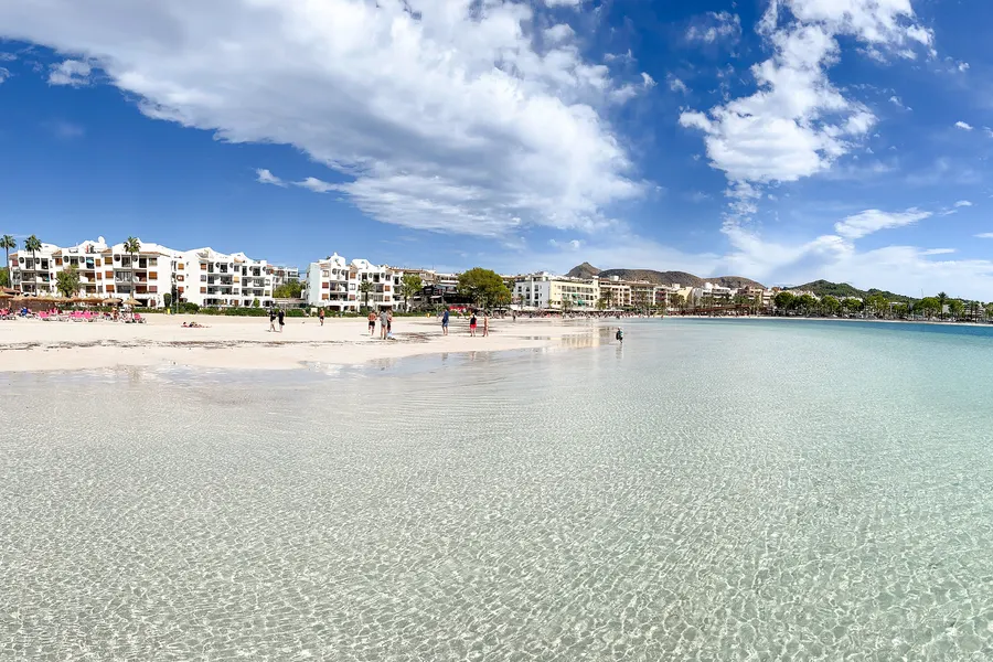 Sehr flaches, glasklares Wasser über hellem Sandgrund im Vordergrund, dahinter eine Sandbank mit einzelnen Badenden und die weißen Apartmenthäuser von Port d'Alcúdia.