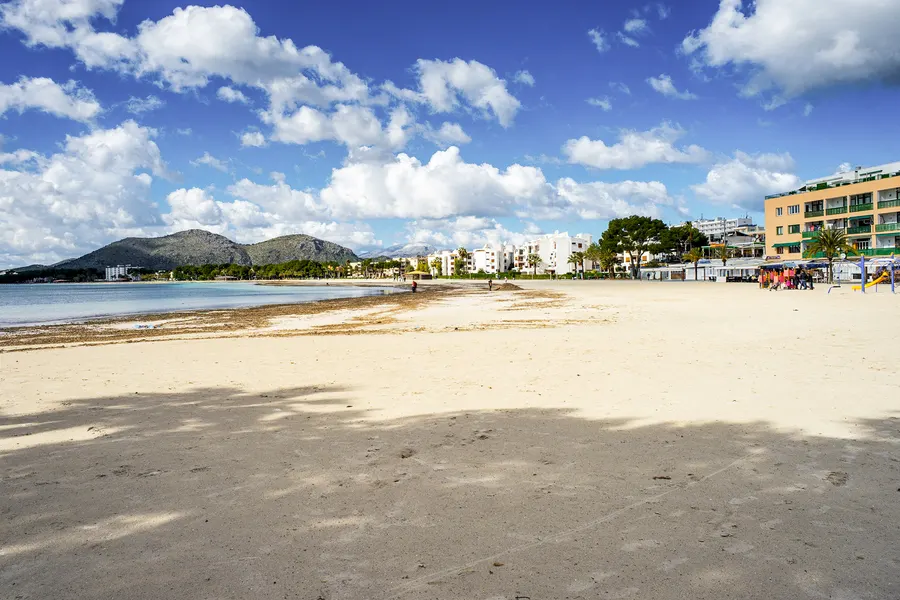 Menschenleerer Strand im Februar mit ungeräumtem Seegrassaum am Wasser, rechts geschlossene Hotelbauten und ein leerer Kinderspielplatz, links die Hügel im Dunst.