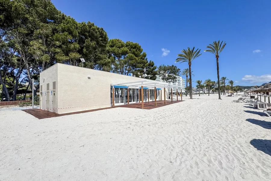 Neu errichteter, heller Balneari-Flachbau mit Holzterrasse und blauen Markisen am Strand, noch mit rot-weißem Absperrband gesichert, dahinter Kiefern und Palmen.