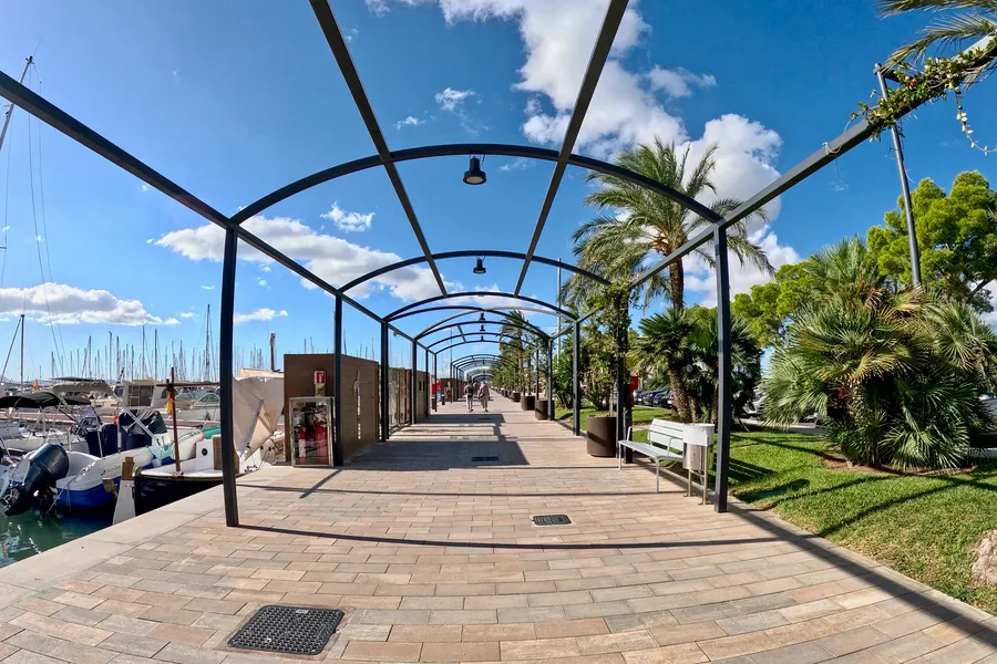 Überdachter Promenadenabschnitt am Sportboothafen mit bogenförmigem Stahlgerüst, links vertäute Boote, rechts Palmen und Rasenflächen, in der Ferne zwei Spaziergänger.
