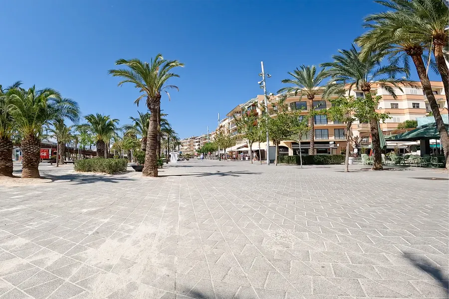 Breite, hell gepflasterte Promenade des Passeig Marítim um die Mittagszeit, gesäumt von Dattelpalmen, rechts Apartmenthäuser mit Läden und Cafés, kaum Menschen unterwegs.
