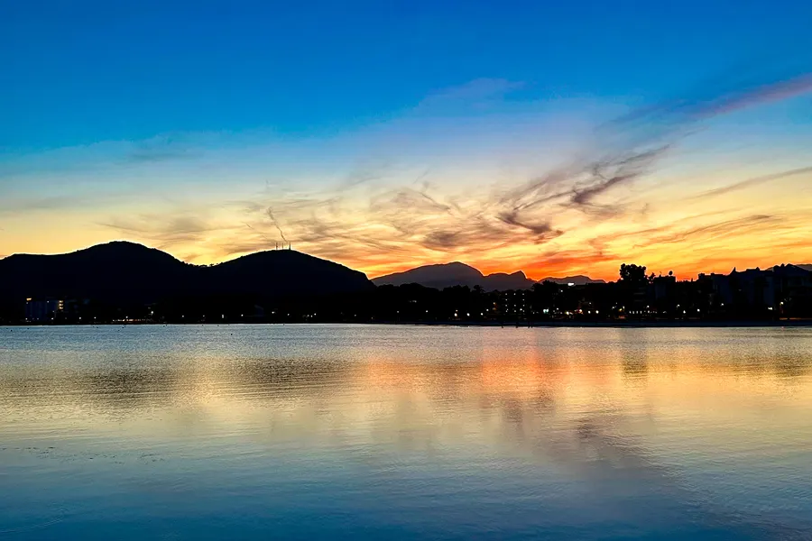 Sonnenuntergang über der Bucht von Alcúdia: orangeroter Himmel mit gefiederten Wolken über der dunklen Silhouette der Tramuntana, davor die Lichter des Orts und das spiegelglatte Wasser.
