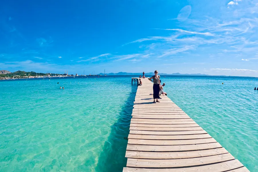 Blick vom Badesteg zurück Richtung Ufer über glasklares, flaches Wasser, am Horizont die Küstenlinie der Bucht und die Berge.

