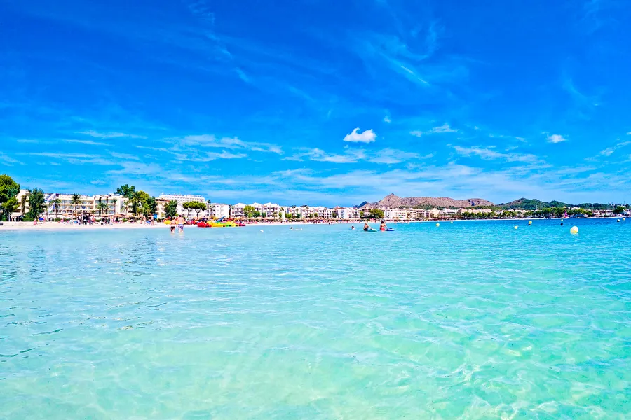 Blick vom Wasser aus auf die Hotelzeile von Port d'Alcúdia hinter dem Strand, davor türkisfarbenes Flachwasser, rechts am Horizont die Berge der Halbinsel Victòria.

