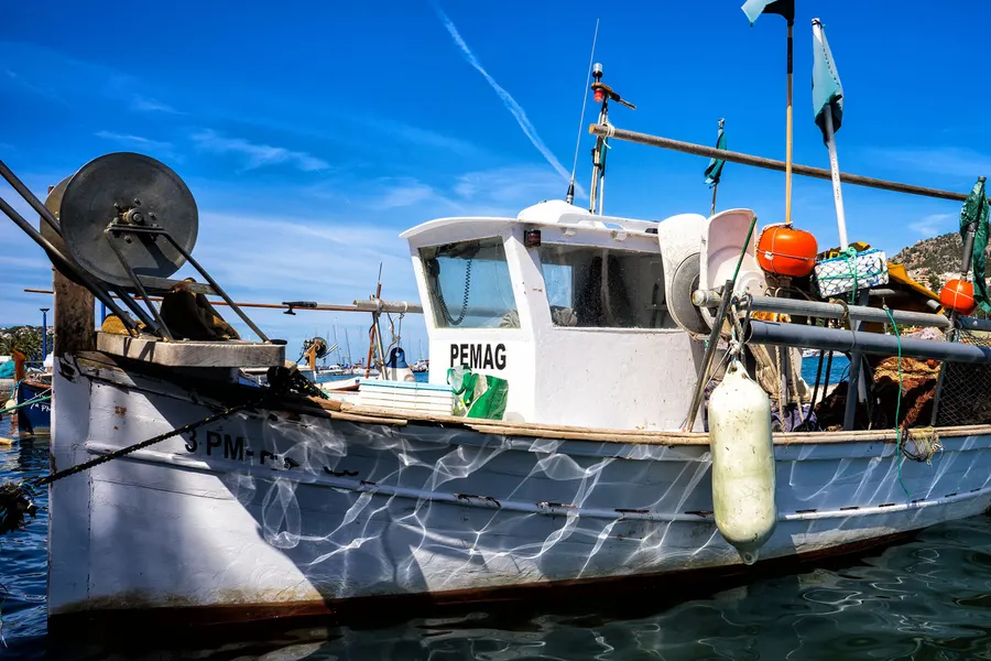 Älteres Fischerboot mit Netzhaspel, Bojen und Fahnen im Hafen von Port d'Andratx, dahinter Masten und Himmel.