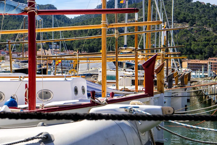 Rot lackierte Holzmasten und Tauwerk eines traditionellen Seglers im Hafen von Port d'Andratx, dahinter weitere Boote und der Hang.