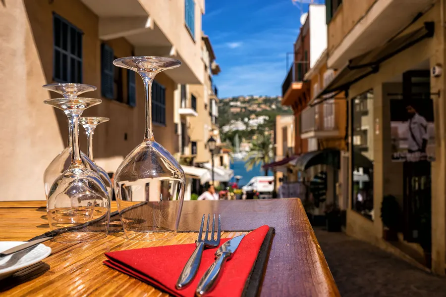 Gedeckter Tisch mit umgedrehten Gläsern und roter Serviette am Rand einer Gasse in Port d'Andratx, im Hintergrund Läden und die Bucht.