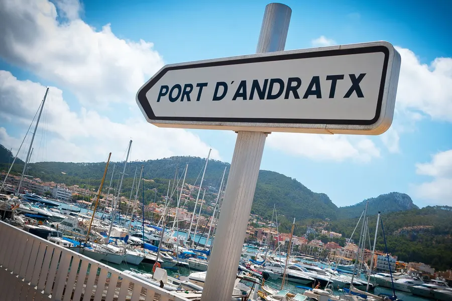 Weißes Straßenschild mit der Aufschrift Port d'Andratx über der Bucht, dahinter Masten, Boote und die bewaldeten Hügel.
