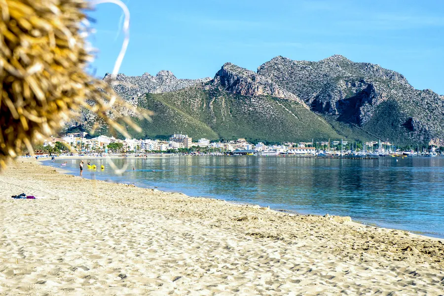 Heller Sandstrand in einer flachen Sichel, dahinter die weiße Häuserzeile des Orts und der Yachthafen, darüber der schroffe Felszug der Serra de Tramuntana; links im Anschnitt ein Strohschirm.
