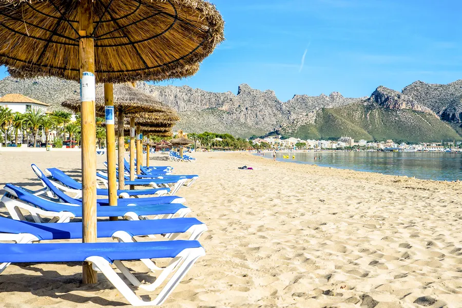 Reihe blau bespannter Strandliegen unter Strohschirmen im Sand, dahinter der flach auslaufende Strand, die Palmen der Promenade und die zerklüftete Felskette im Hintergrund.
