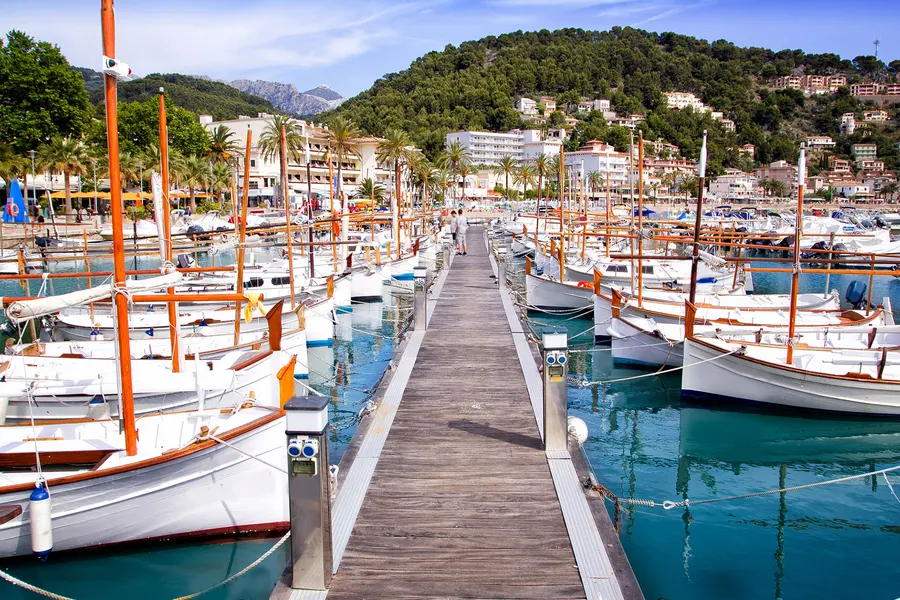 Hölzerner Steg zwischen zwei Reihen weißer Llaüts mit hohen Masten im Hafen von Port de Sóller, dahinter der bewaldete Hang.