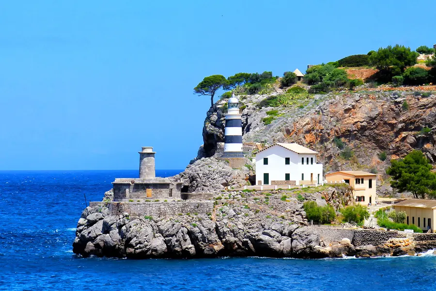Weiß-schwarz geringelter Leuchtturm auf einem Felsvorsprung an der Einfahrt von Port de Sóller, daneben ein alter Wachturm und tiefblaues Meer.