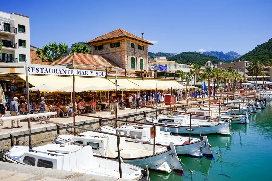 Promenade von Port de Sóller mit einem Restaurant unter Markisen, Passanten und Booten im Vordergrund, dahinter die Berge.