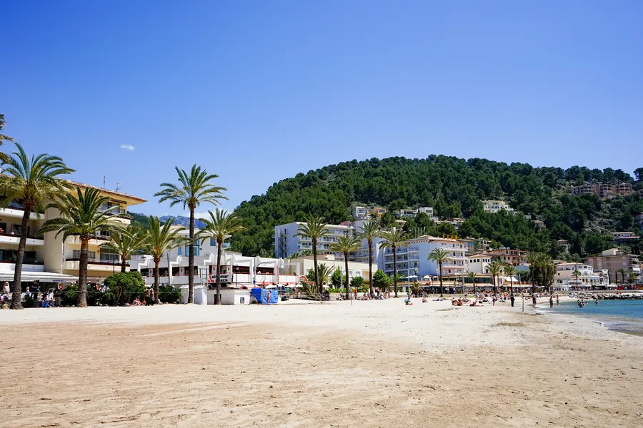 Breiter Sandstrand von Port de Sóller mit Palmen an der Promenade, Badegästen und dem bewaldeten Hang dahinter.

