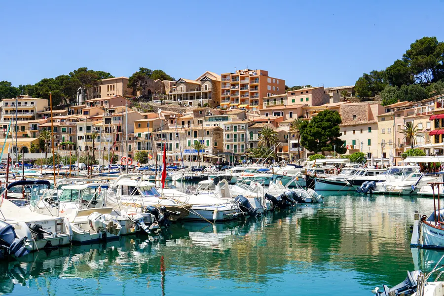 Dicht belegter Yachthafen von Port de Sóller mit Motorbooten, dahinter die Häuserzeile am Hang und Kiefern.
