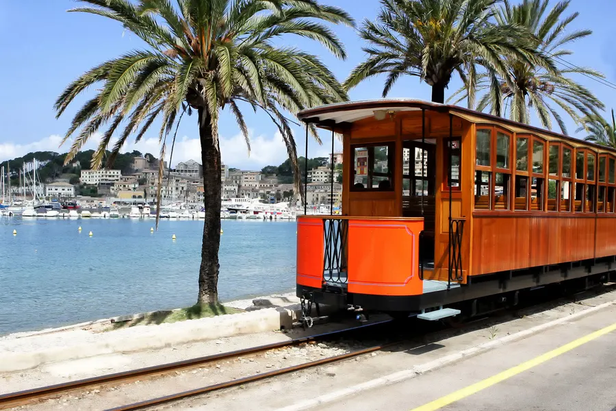 Die orangefarbene Straßenbahn von Sóller auf ihrem Gleis an der Promenade von Port de Sóller, dahinter Palmen und die Bucht.
