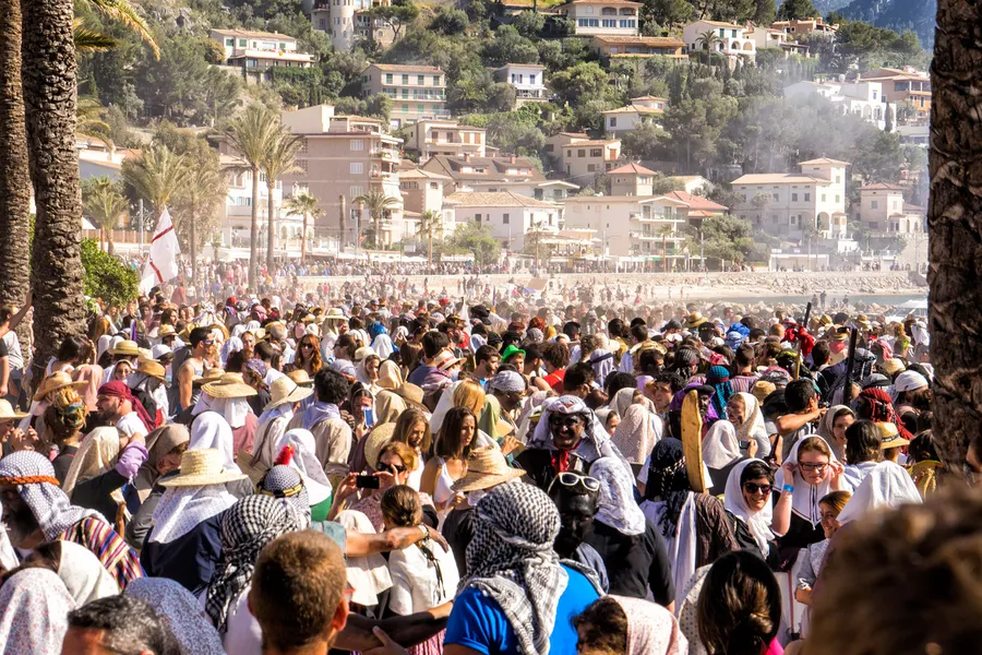Dichte Menschenmenge in Kostümen am Strand von Port de Sóller während des Fests Es Firó, dahinter der Ort am Hang.
