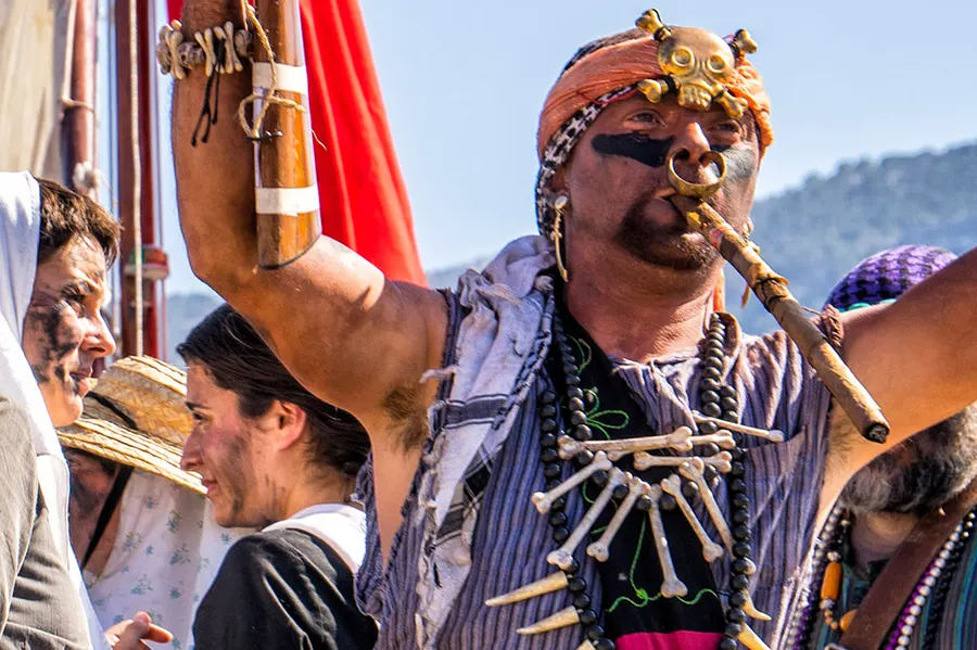 Kostümierter Teilnehmer mit Halskette, Kopfschmuck und erhobenem Arm beim Fest Es Firó, umgeben von weiteren Teilnehmern.
