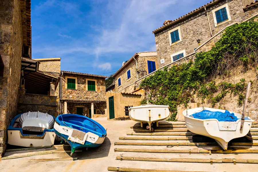 Mehrere kleine Ruderboote auf hölzernen Rampen vor den Natursteinhäusern von Port de Valldemossa, dahinter der bewachsene Steilhang.