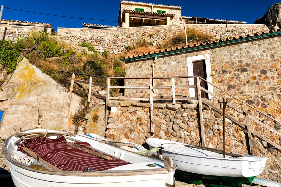 Bootshaus aus Bruchstein mit Steintreppe und Geländer in Port de Valldemossa, davor zwei weiße Boote mit rot gestreiften Planen.