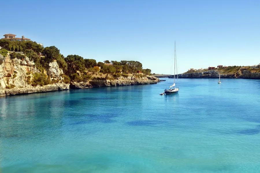 Der Strand von Porto Cristo mit Schirmen und Badegästen, dahinter die mehrgeschossige Hotel- und Wohnbebauung am Hang.