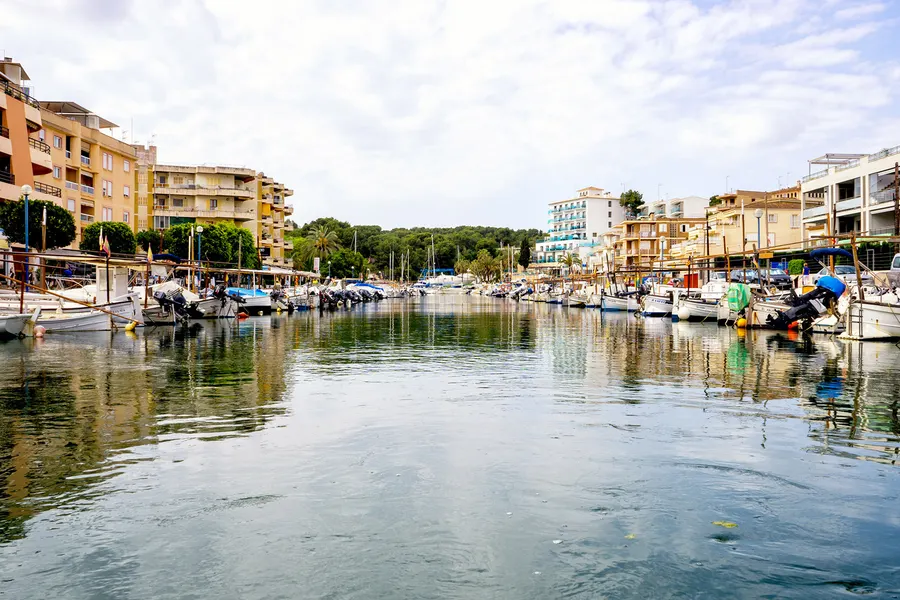 Ruhiges Hafenbecken von Porto Cristo mit Booten an beiden Ufern und der Bebauung, die sich im Wasser spiegelt.
