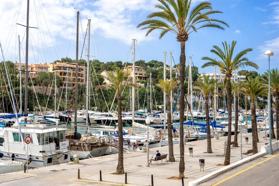 Sportboothafen von Porto Cristo mit Masten, Stegen und einer Reihe hoher Palmen entlang der Uferpromenade.
