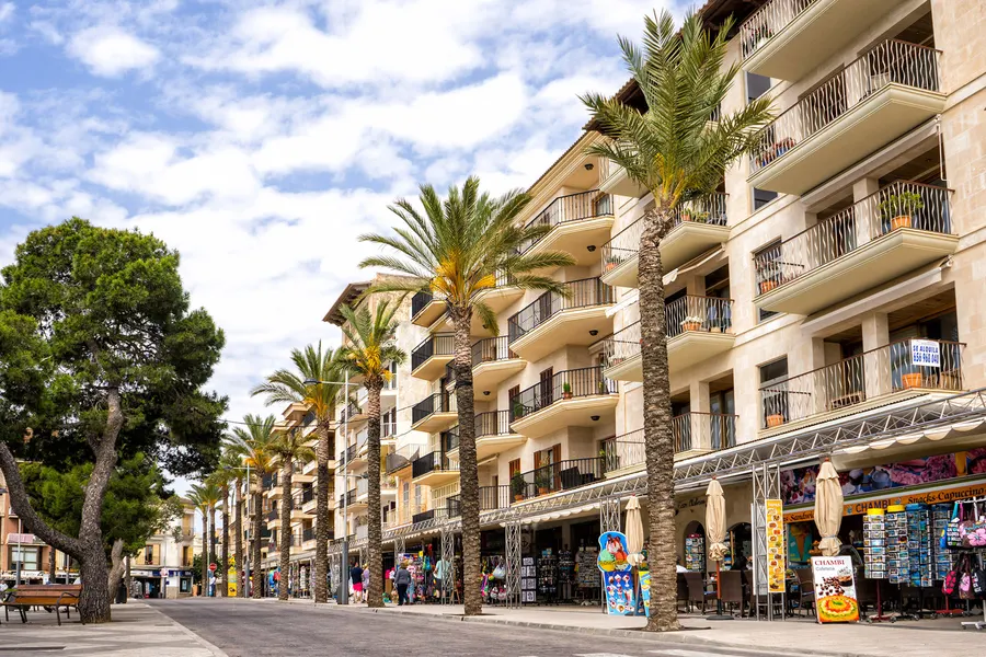 Uferpromenade von Porto Cristo mit Palmenreihe, parkenden Autos und Wohnhäusern mit Balkonen unter blauem Himmel.
