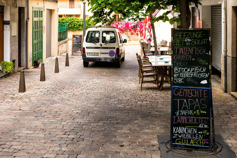Gepflasterte Gasse in Porto Cristo mit einem Lokal, Tafelmenü und Sonnenschirmen, dahinter Bäume und niedrige Häuser.
